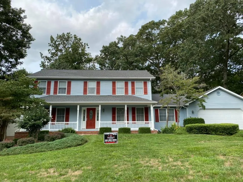 Light blue two-story colonial in Waldorf, MD with red shutters, wraparound porch and Summit Exteriors Storm Damage yard sign after a roof replacement