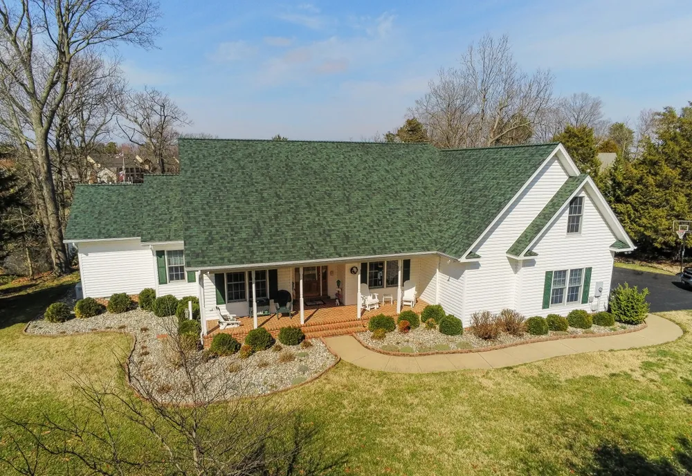 Aerial photo of a white farmhouse-style home in St. Leonard, MD with a new green architectural shingle roof and wraparound porch