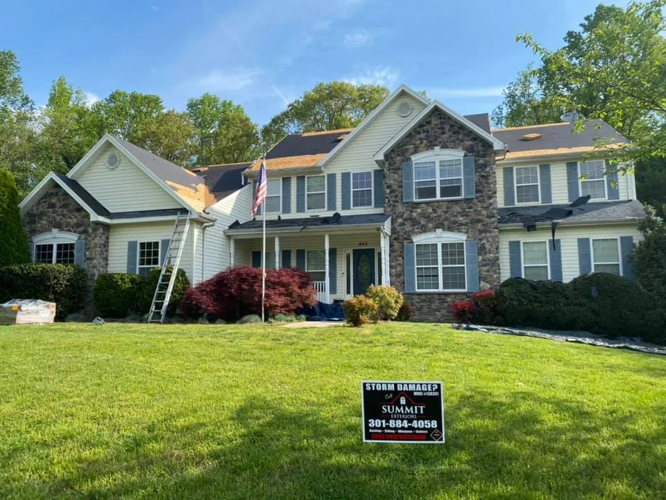 Two-story stone-front colonial in Huntingtown, MD with Summit Exteriors crew installing new architectural shingles and a Storm Damage yard sign in front