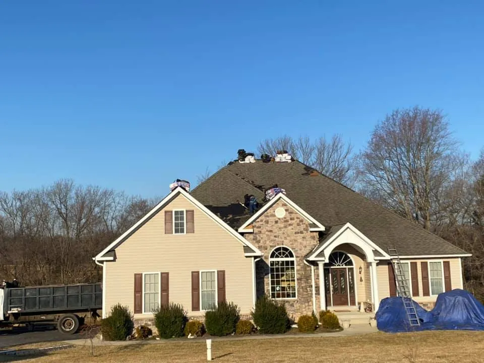 Stone-front rancher in Prince Frederick, MD with Summit Exteriors crew on the steep hip roof installing new architectural shingles and a dump trailer below