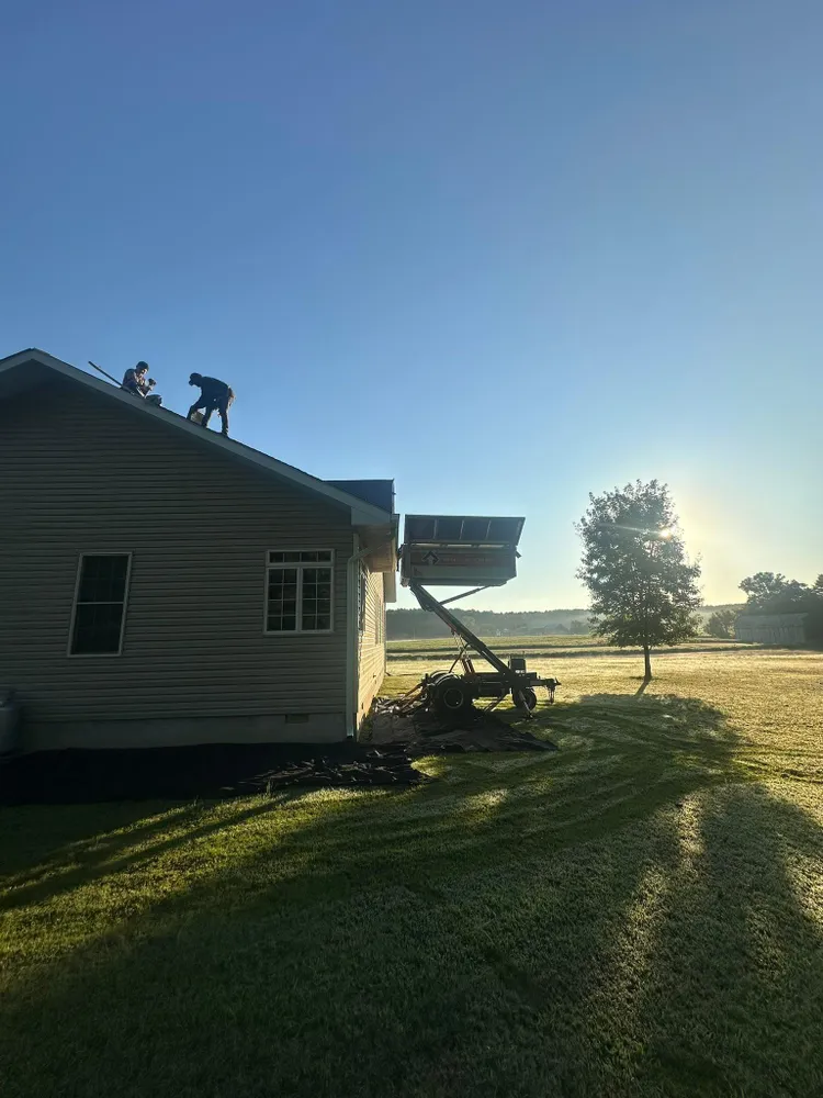 Summit Exteriors crew on the roof at sunrise tearing off old shingles with a material lift trailer beside a Mechanicsville, MD home, completed as part of a roof replacement job in Mechanicsville, MD
